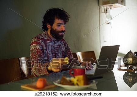 Stock Image - Bearded young man having breakfast, using laptop at home. Fotosearch