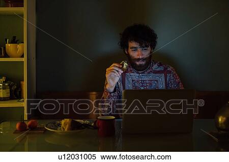 Stock Photography - Bearded young man having breakfast, using laptop at home. Fotosearch