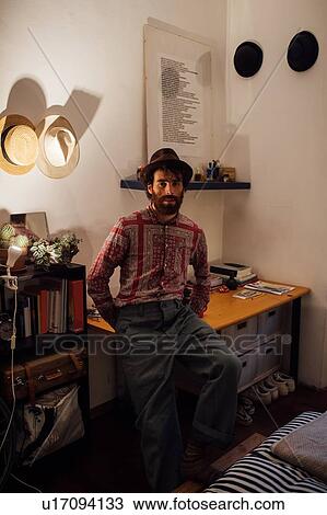 Stock Image - Bearded young man sitting on bedroom table. Fotosearch