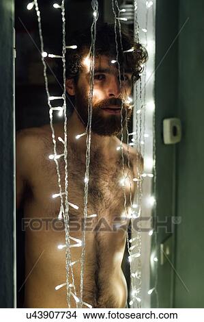 Picture - Bearded young man standing behind string of decorative lights. Fotosearch