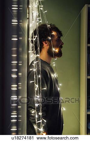 Stock Image - Bearded young man standing between string of decorative lights. Fotosearch