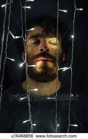 Stock Photograph - Bearded young man standing between string of decorative lights. Fotosearch