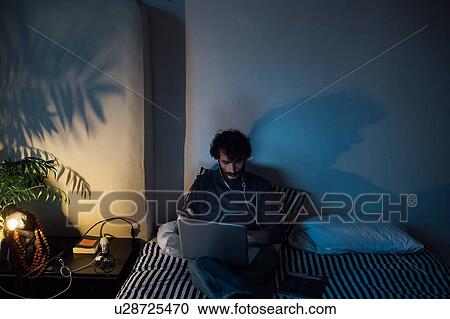 Stock Image - Bearded young man using laptop in bed. Fotosearch