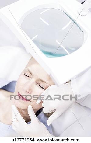 Picture - Beauty technician examining woman's facial skin using magnifying glass, close-up.. Fotosearch
