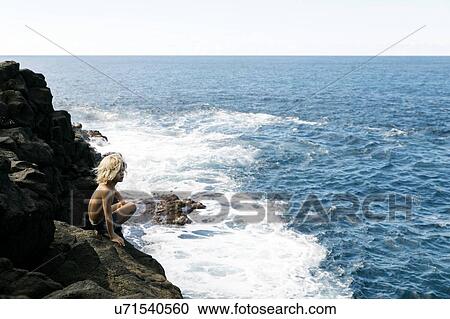 Stock Image - Boy crouching on cliff by sea. Fotosearch