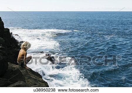Stock Photo - Boy crouching on cliff by sea. Fotosearch