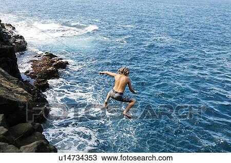 Stock Image - Boy jumping off cliff by sea. Fotosearch