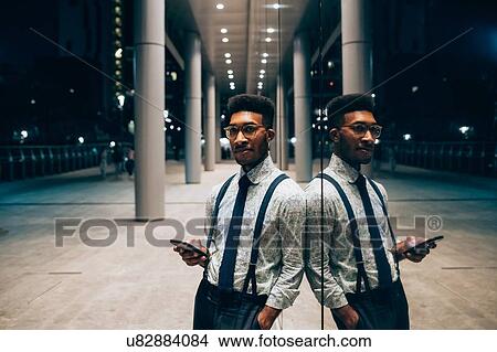 Picture - Businessman using smartphone, leaning against mirrored wall of office building, Milano, Lombardia, Italy. Fotosearch