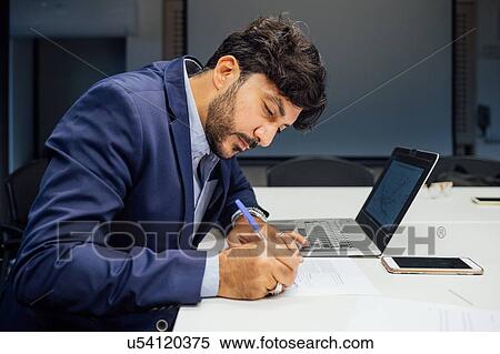 Stock Photography - Businessman writing notes and using laptop in office. Fotosearch