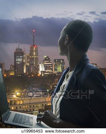 Stock Image - Businesswoman holding laptop by city skyline at sunset in Nairobi, Kenya. Fotosearch