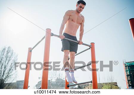 Picture - Calisthenics at outdoor gym, bare chested young man doing push up on exercise equipment. Fotosearch
