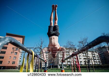 Stock Image - Calisthenics at outdoor gym, bare chested young man doing handstand on parallel bars, low angle view. Fotosearch