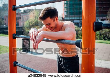 Stock Photo - Calisthenics at outdoor gym, bare chested young man leaning on exercise equipment exhausted. Fotosearch