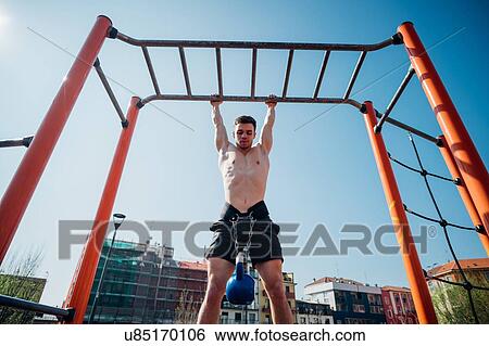 Stock Photograph - Calisthenics at outdoor gym, bare chested young man hanging from exercise equipment with kettlebell on waist harness. Fotosearch