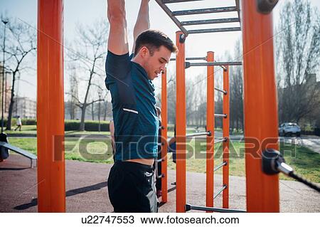 Stock Image - Calisthenics at outdoor gym, young man hanging from exercise equipment. Fotosearch