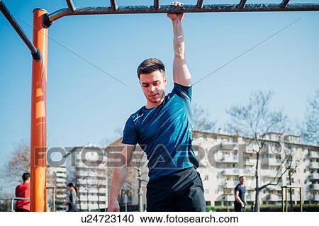 Stock Image - Calisthenics at outdoor gym, young man hanging by one arm from exercise equipment. Fotosearch