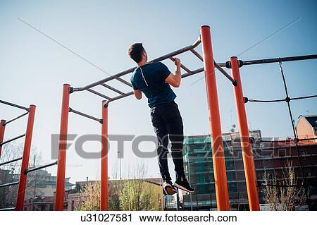 Stock Image - Calisthenics at outdoor gym, young man doing pull ups on exercise equipment, rear view. Fotosearch