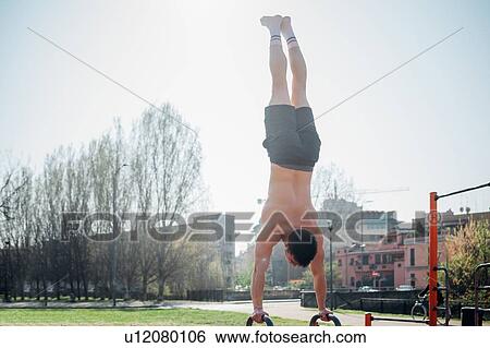 Stock Photograph - Calisthenics at outdoor gym, young man doing handstand on exercise equipment, rear view. Fotosearch
