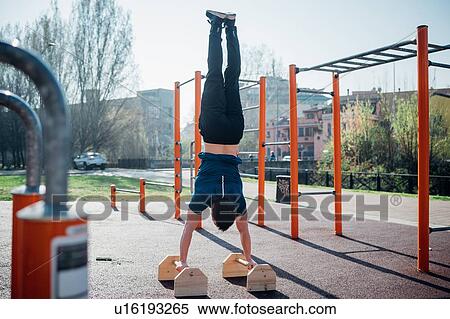 Stock Photography - Calisthenics at outdoor gym, young man doing hand stand on exercise equipment, rear view. Fotosearch