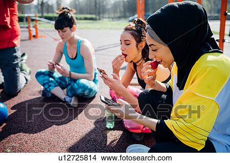 Picture - Calisthenics class at outdoor gym, young women sitting looking at smartphones and eating fruit. Fotosearch