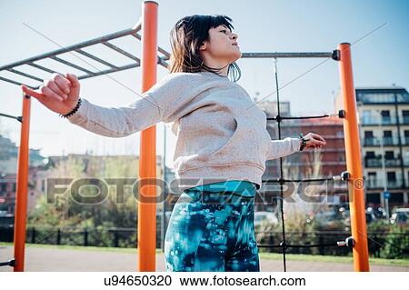 Stock Image - Calisthenics class at outdoor gym, young woman jumping with arms outstretched. Fotosearch