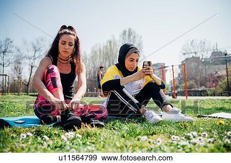 Stock Photo - Calisthenics class at outdoor gym, young women sitting on grass tying laces and looking at smartphone. Fotosearch