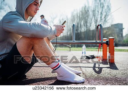 Stock Photograph - Calisthenics class at outdoor gym, young man sitting down and looking at smartphone. Fotosearch
