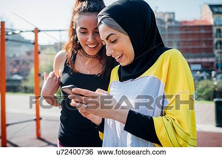 Stock Photograph - Calisthenics class at outdoor gym, two young women looking at smartphone. Fotosearch