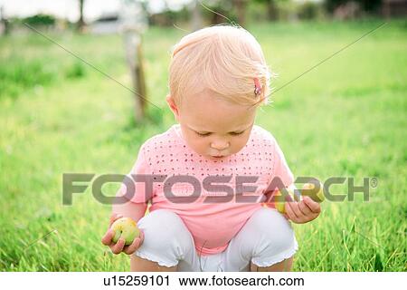 Stock Image - Cute female toddler squatting on grass looking down. Fotosearch