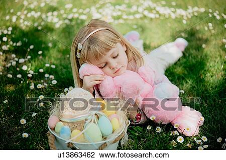 Stock Image - Cute girl lying on grass with toy rabbit and easter egg basket. Fotosearch