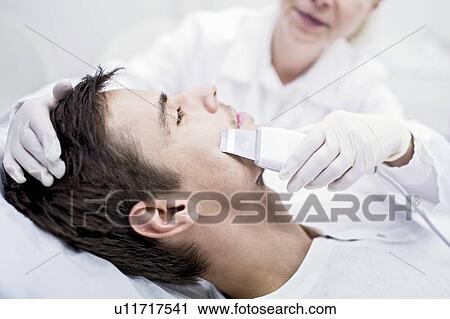 Stock Image - Dermatologist applying facial microdermabrasion treatment on man in clinic, close-up. The cosmetic procedure uses micro crystals to remove dead skin cells. This exfoliating treatment can stimulate the production of collagen and help improve fine lines.... Fotosearch