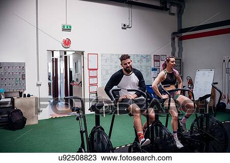 Stock Image - Exhausted young woman and man training together on gym exercise bikes, taking a break. Fotosearch