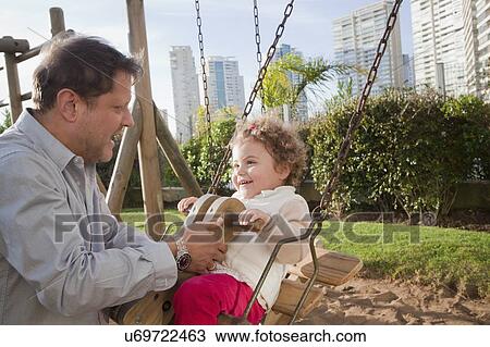 Stock Image - Father by daughter on swing in city park. Fotosearch