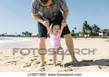 Stock Image - Father helping his baby girl walk on beach. Fotosearch