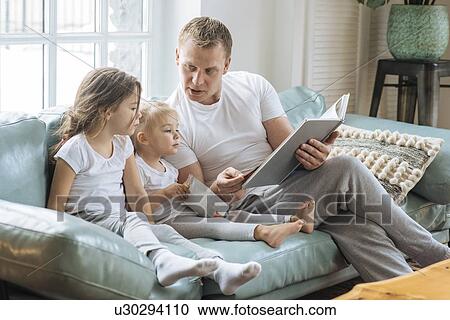 Stock Image - Father reading book to daughters on sofa. Fotosearch