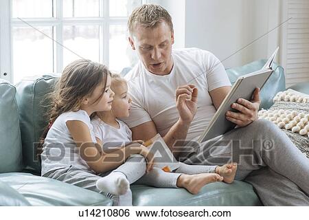 Stock Photograph - Father reading book to daughters on sofa. Fotosearch