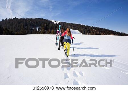Stock Image - Five teenage girl skiers carrying skis in snow covered landscape, rear view, Tyrol, Styria, Austria. Fotosearch