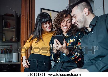 Stock Image - Friends using smartphone at home. Fotosearch