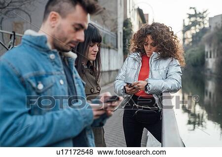 Stock Photo - Friends using smartphone by river, Milano, Lombardia, Italy. Fotosearch