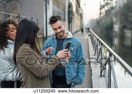 Friends using smartphone by river, Milano, Lombardia, Italy View Large Photo Image Stock Photography - Friends using smartphone by river, Milano, Lombardia, Italy. Fotosearch