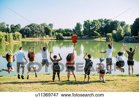 Stock Photo - Group of friends jumping in front of lake in park. Fotosearch