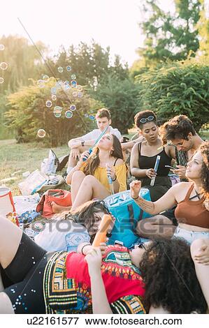 Stock Photo - Group of friends relaxing, blowing bubbles at picnic in park. Fotosearch