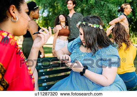 Stock Image - Group of friends relaxing, using smartphone in park. Fotosearch