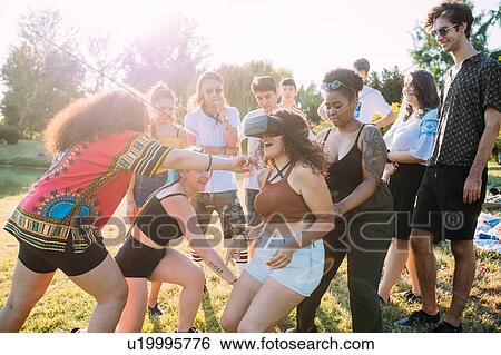 Stock Photograph - Group of friends teasing woman looking into VR headset in park. Fotosearch