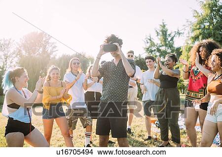 Picture - Group of friends watching man looking into VR headset in park. Fotosearch