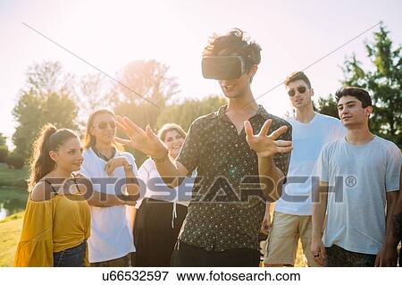 Stock Photo - Group of friends watching man looking into VR headset in park. Fotosearch