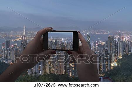 Stock Image - Hands of woman taking photograph with smart phone of cityscape in Hong Kong, China. Fotosearch