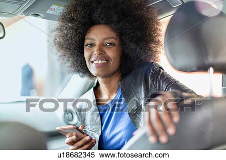 Stock Photography - Happy young woman with afro hair in car passenger seat holding smartphone, portrait. Fotosearch
