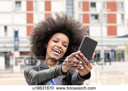 Stock Image - Happy young woman with afro hair in city, taking smartphone selfie. Fotosearch