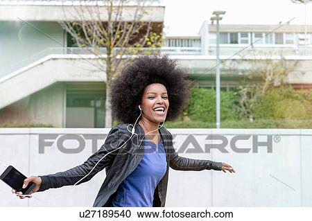 Stock Image - Happy young woman with afro hair in city, laughing and dancing to smartphone music. Fotosearch
